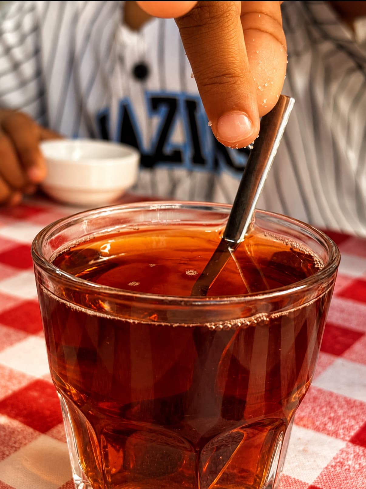 A hand stirring a small glass of amber tea beside a saucer of sugar